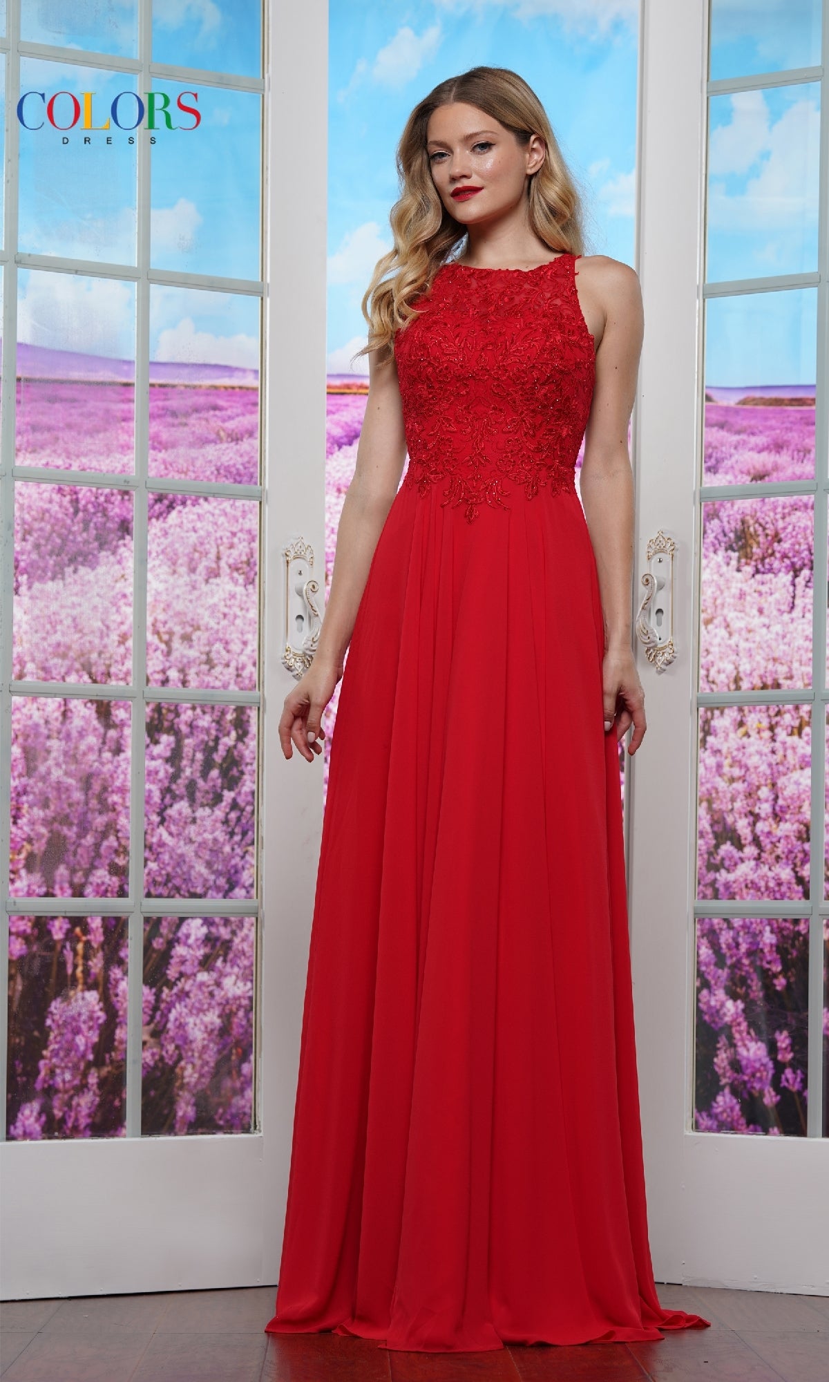 A woman stands indoors before glass doors, wearing the Colors Dress 3440—a sleeveless, high-neck, floor-length red prom dress with lace details. Purple flower fields are visible through the doors behind her.